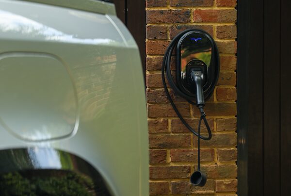 A wall-mounted black EV home charger with coiled cable on a brick wall beside a white car