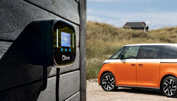 An Ohme home EV charger mounted on a dark wooden wall with an orange and white VW ID. Buzz electric van parked nearby