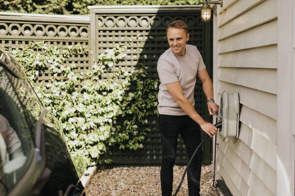 A man plugging a charging cable into a wall-mounted home EV charge point beside a parked car in a garden driveway