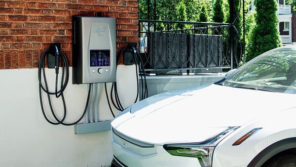 A dcbel home energy station EV charger mounted on a brick wall with cables, next to a white electric car