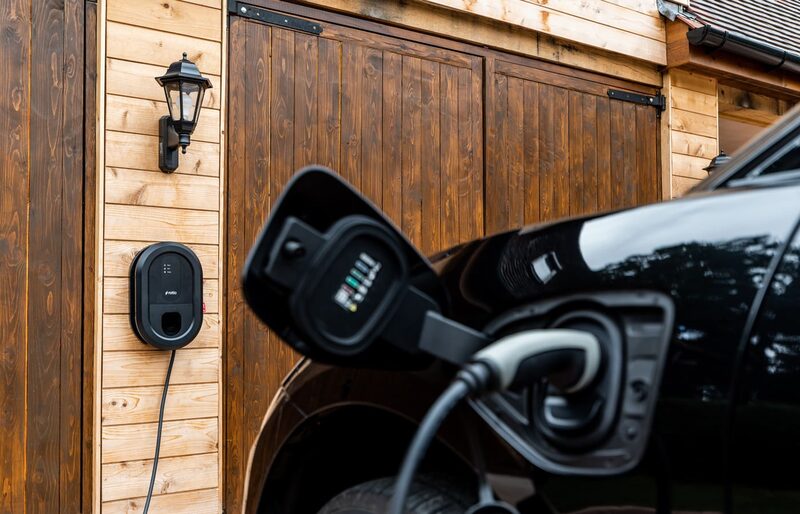 A black Ratio EV wall charger mounted on a timber garage wall beside a black car with its charging port open and cable plugged in