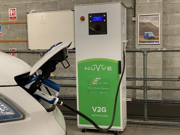 A Nuvve e-flex V2G bidirectional EV charger in an industrial building with a charging cable plugged into a white electric car