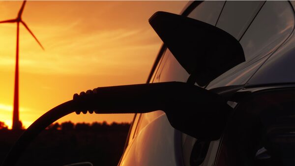 An electric car charging cable plugged into a car at sunset with a wind turbine in the background