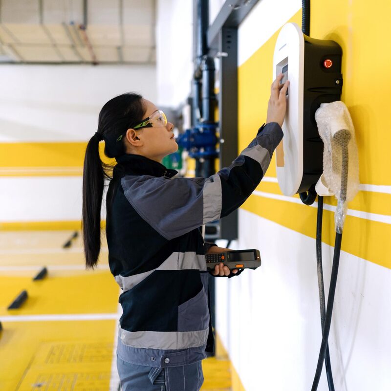 A technician in safety glasses testing a wall-mounted EV charge point in a yellow-marked car park