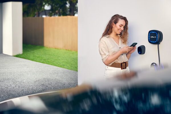 A woman using her phone next to a Wallbox Pulsar Max EV charger mounted on a wall outside a house
