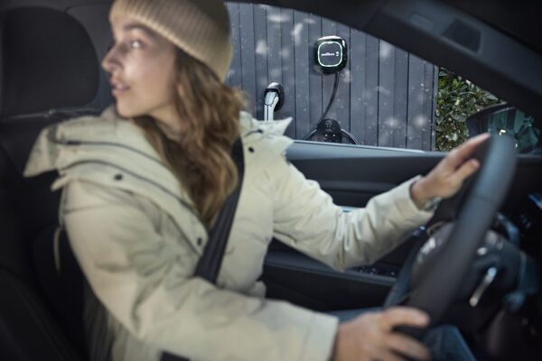 A woman driving an electric car parked outside a dark timber building with a Wallbox EV charger mounted on the wall