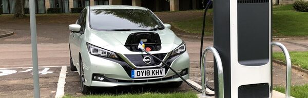 A pale green Nissan Leaf plugged into a public charge point in a car park