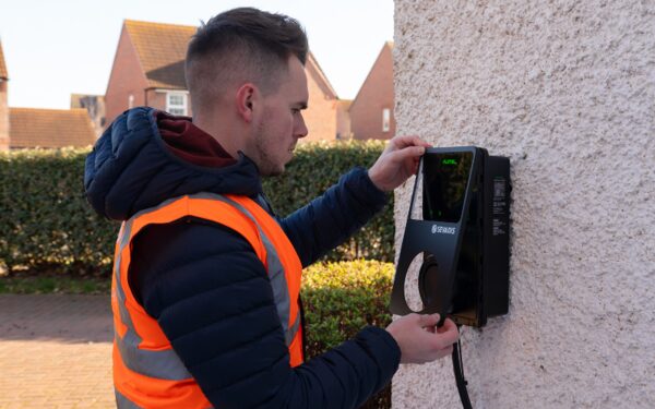An installer in a high-visibility vest fitting a black Sevadis Maxicharger EV chargepoint to an outside house wall