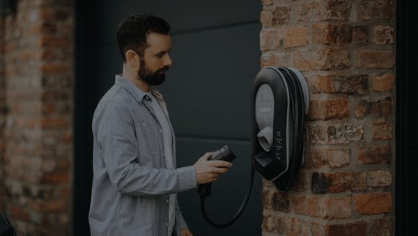 A man holding the charging plug of a black zappi wall-mounted EV charger installed on a brick wall outside a garage