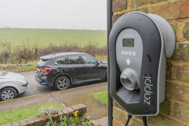 A myenergi zappi EV charger mounted on a brick house wall with an electric car charging on a driveway in the background