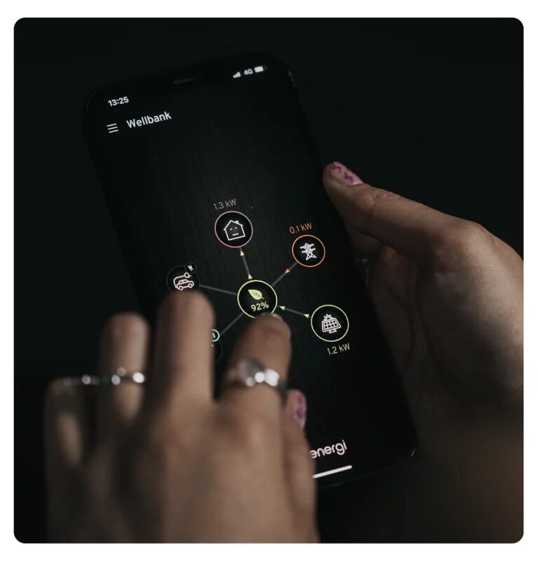 A person's hands holding a smartphone showing the myenergi app with an energy flow diagram between home, grid, EV charger and solar icons