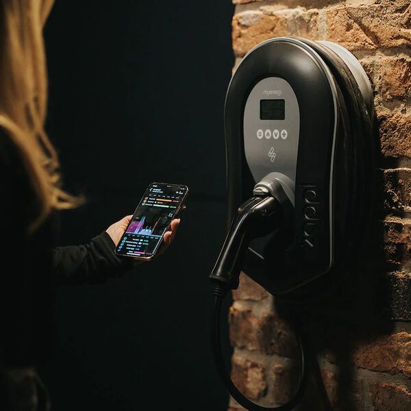 A woman holding a smartphone next to a black myenergi zappi EV charger mounted on a brick wall