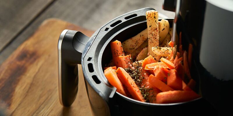 A close-up of an air fryer basket filled with seasoned carrots and parsnips on a wooden board