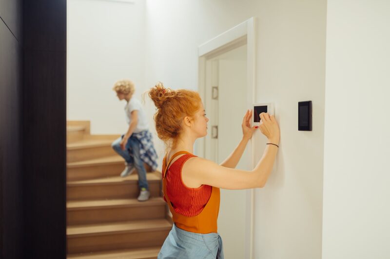 A woman adjusting a wall-mounted heating thermostat in a home hallway while a child walks down the stairs behind her