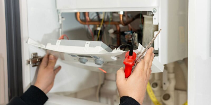 A heating engineer's hands removing the front casing of a wall-mounted gas boiler with a screwdriver, exposing internal pipework and wiring