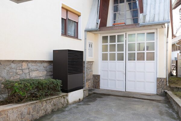 An outdoor air source heat pump unit installed against the exterior wall of a house beside garage doors