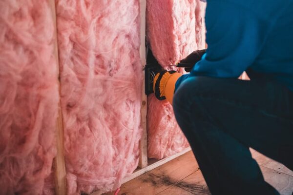 A worker in a blue jacket fitting pink fibreglass insulation batts between timber wall studs