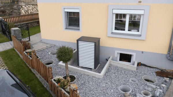 An outdoor heat pump unit installed on a gravel bed against the exterior wall of a house