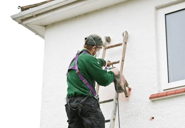 A worker on a ladder drilling holes in the external wall of a white house to install cavity wall insulation