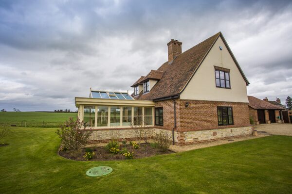 A large brick-and-render country house with a glazed conservatory extension, lawn and fields under a cloudy sky, with a green LPG tank cover in the garden