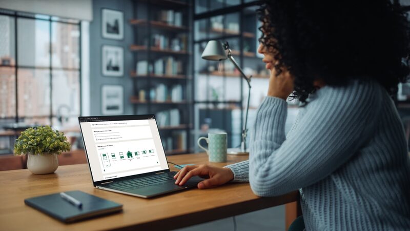 A woman at a desk using a laptop showing a home energy efficiency tool with house icons