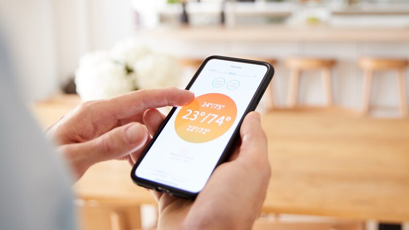 A person using a smartphone app to control a smart thermostat showing 23°/74° in a home kitchen