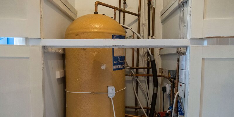 A yellow insulated hot water cylinder with copper pipework installed in an airing cupboard