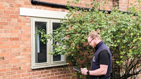 An assessor in a branded gilet taking notes on a clipboard outside a brick house beside a window and shrubs