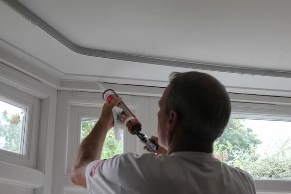 A man using a sealant gun to apply draught-proofing sealant along the ceiling edge above a bay window