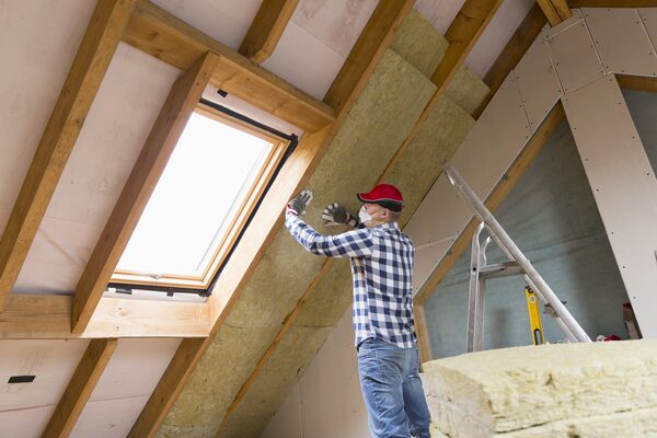 A worker in a red cap fitting mineral wool insulation between roof rafters in a loft conversion beside a skylight