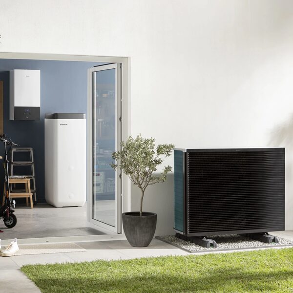 A Daikin heat pump outdoor unit beside a house wall with an indoor cylinder unit visible through an open door