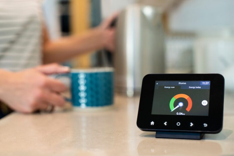 A smart energy monitor on a kitchen worktop showing live electricity usage, with a person making tea in the background
