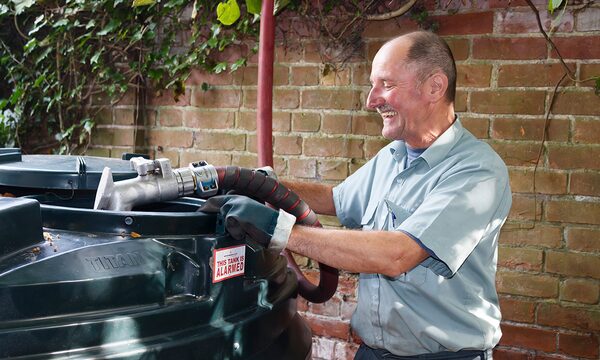 A smiling delivery driver filling a green Titan heating oil tank with a hose against a brick wall