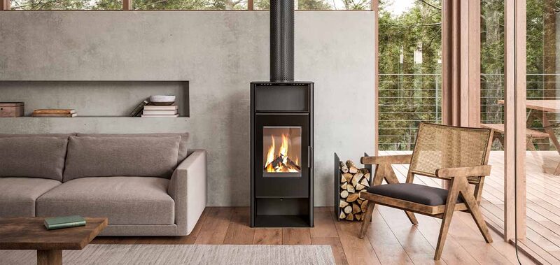 A black freestanding wood-burning stove with a glass door showing flames, installed in a modern living room with a sofa, armchair and a log basket beside it