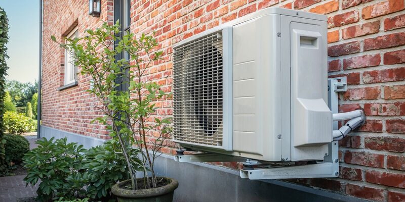 An air source heat pump unit mounted on the outside brick wall of a house next to a potted shrub