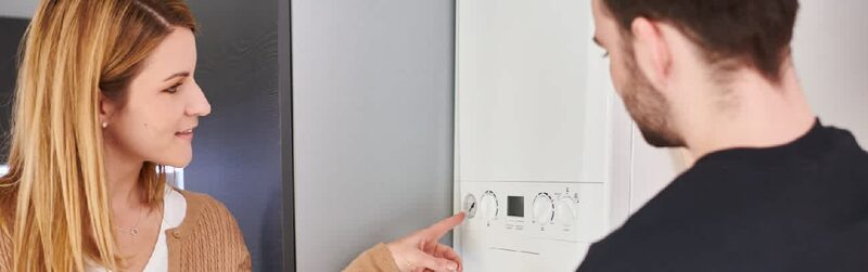 A woman pointing at the controls of a wall-mounted white combi boiler while a man looks on