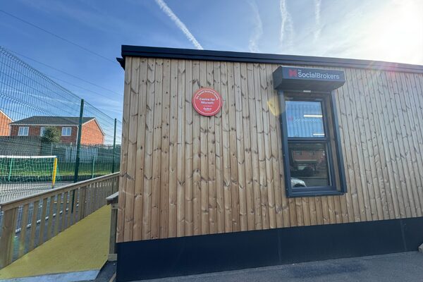 A wooden-clad community building with a red 'Centre for Warmth' plaque and a SocialBrokers sign above the window