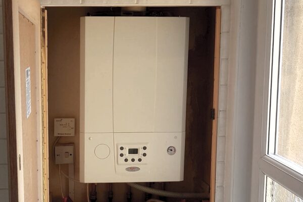 A white wall-mounted combi gas boiler installed in an airing cupboard next to a window, with a heating control panel beside it