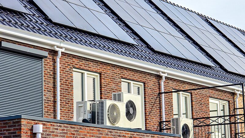 Air source heat pump units mounted on the wall of a brick house with solar panels on the tiled roof