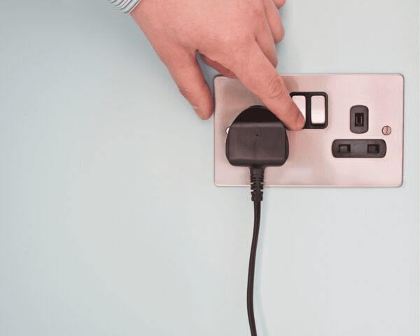 A hand switching off a UK wall socket with a black plug and cable plugged in