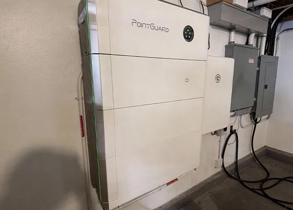 A white PointGuard home battery unit mounted on a garage wall next to electrical panels and cabling