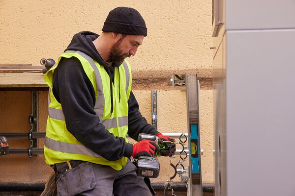 An installer in a hi-vis vest using a cordless drill while working on an outdoor heat pump unit