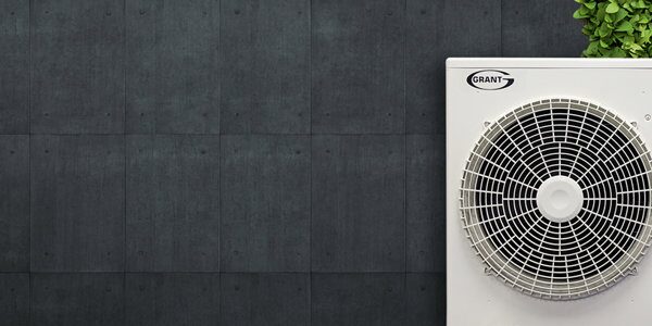 A white Grant air source heat pump outdoor unit with fan grille against a dark grey tiled wall with a green plant