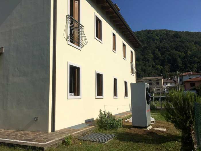 An air source heat pump unit installed on the grass beside a pale yellow multi-storey residential building with hills behind