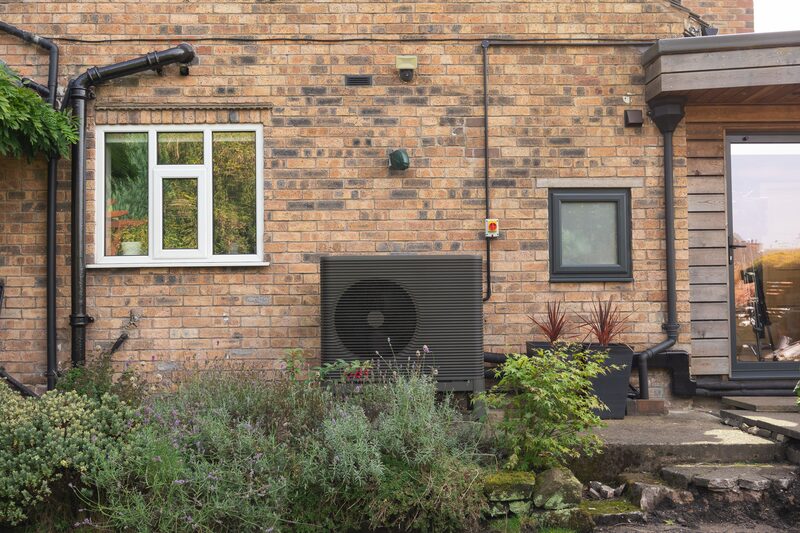 An air source heat pump unit installed on the exterior brick wall of a house beside a window and garden plants