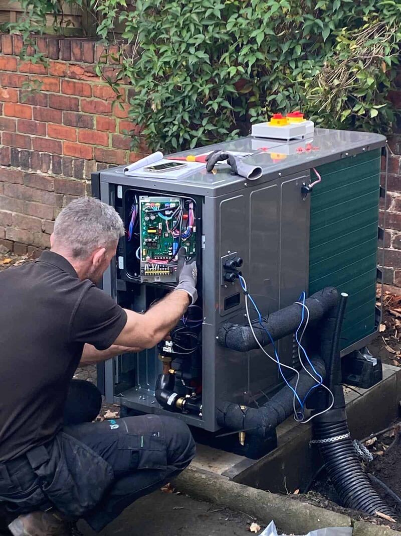 A technician working on the open control panel of an air source heat pump unit installed outside a brick house