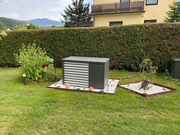 An outdoor grey heat pump unit installed in a garden on a gravel bed beside a hedge
