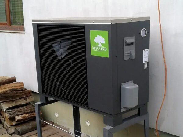 A Mycond air source heat pump unit installed outside a house on a metal stand, next to a stack of firewood