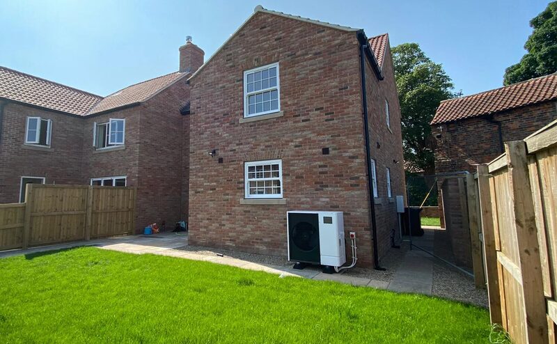 An air source heat pump unit installed on the exterior brick wall of a modern UK house with a lawn and wooden fence
