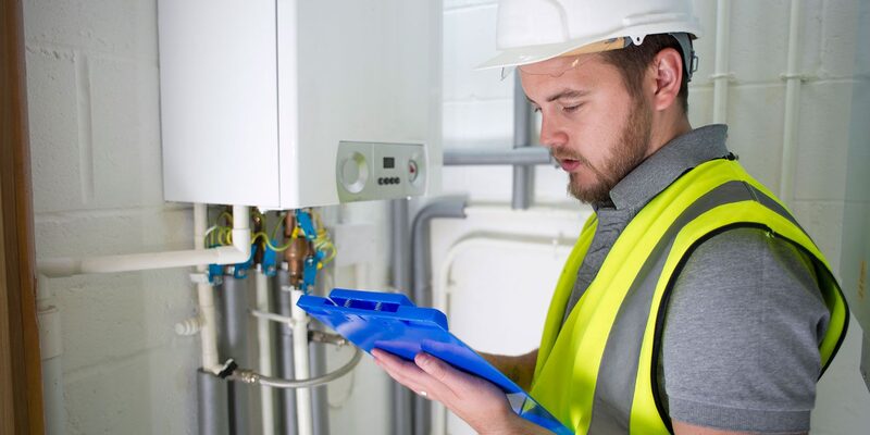 A heating engineer in a hi-vis vest and hard hat checks a clipboard next to a wall-mounted gas boiler and pipework